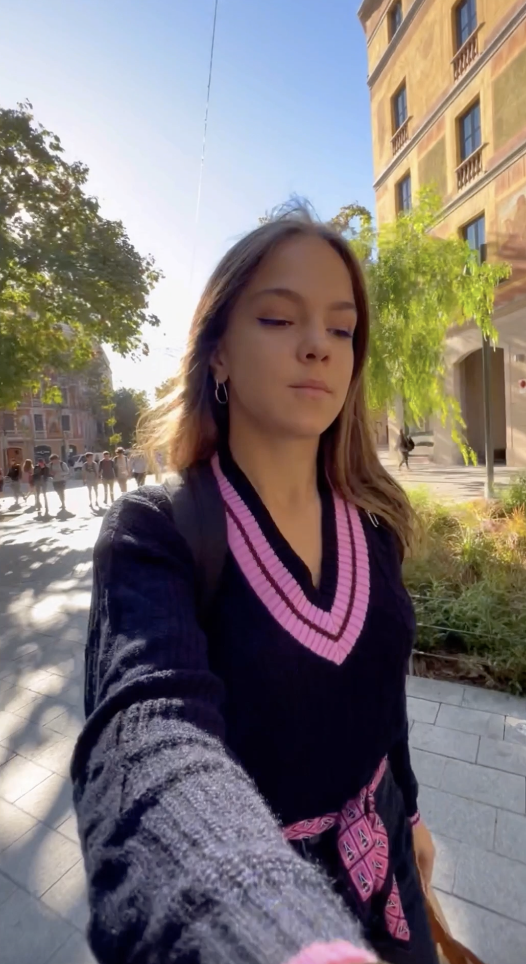 A person with long hair walks along a sunlit street, wearing a black sweater with a pink V-neck. Trees and a building are in the background, with a group of people in the distance.